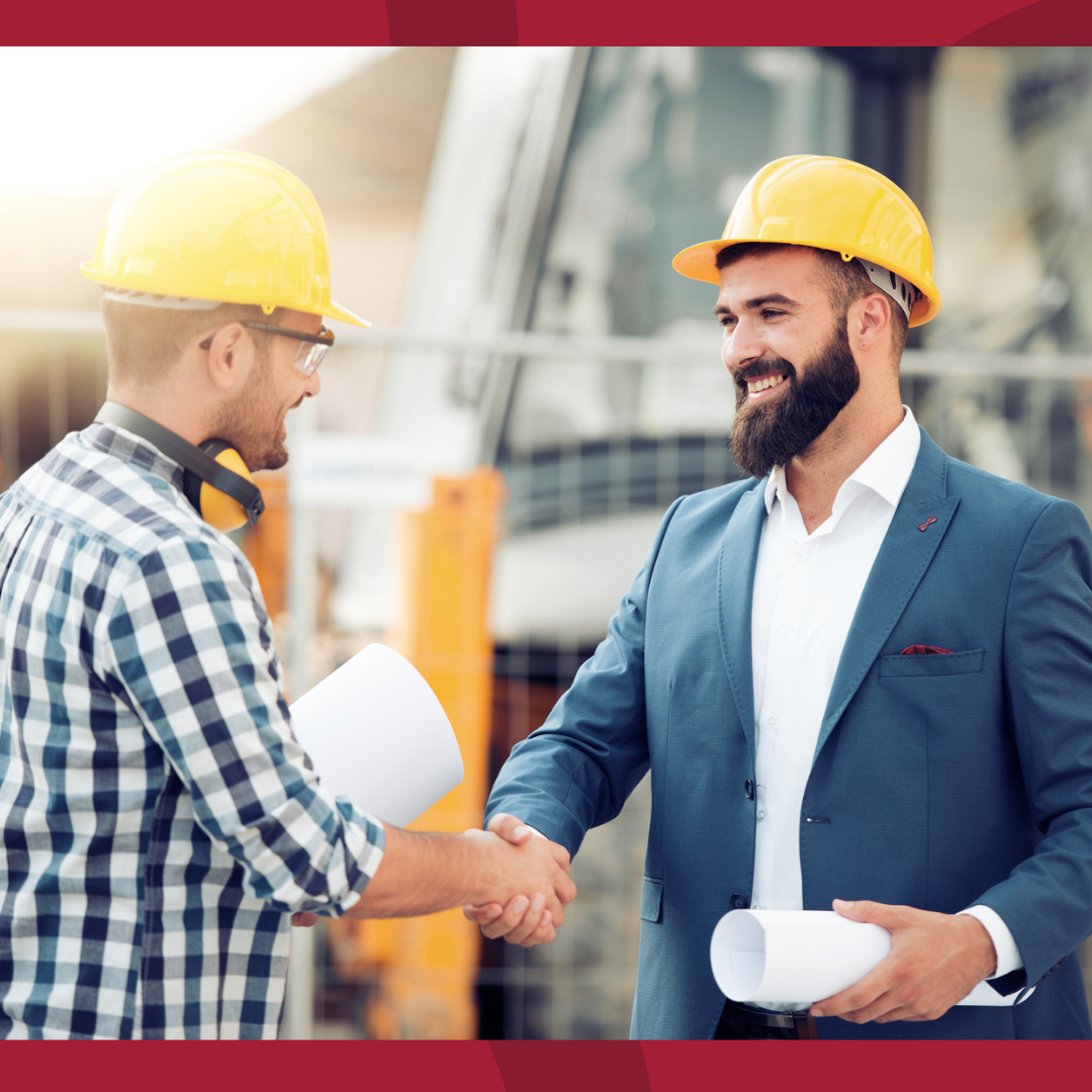 A business man and a construction worker, both in hard hats, are shaking handings on a work site.